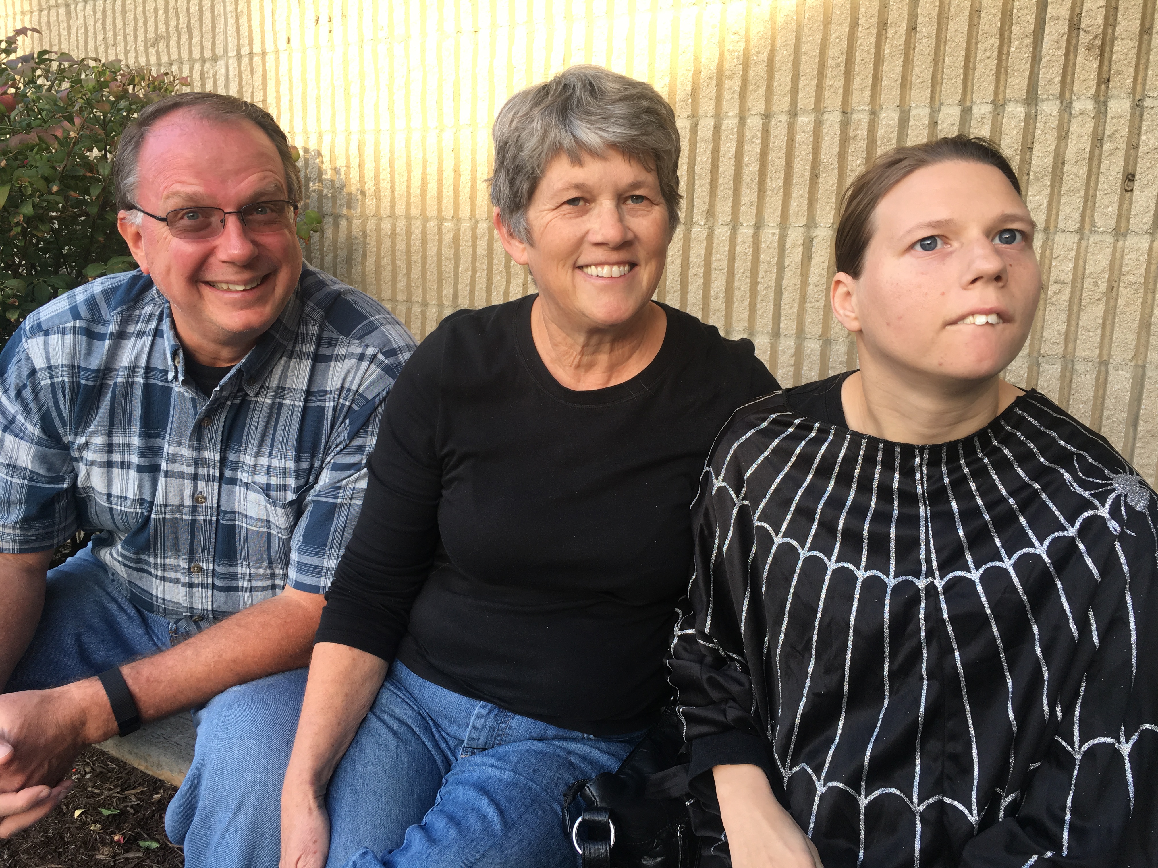 Smiling family sitting together outdoors — a man in a plaid shirt, a woman in a black top, and a young woman in a black spiderweb-patterned outfit, sitting against a brick wall.