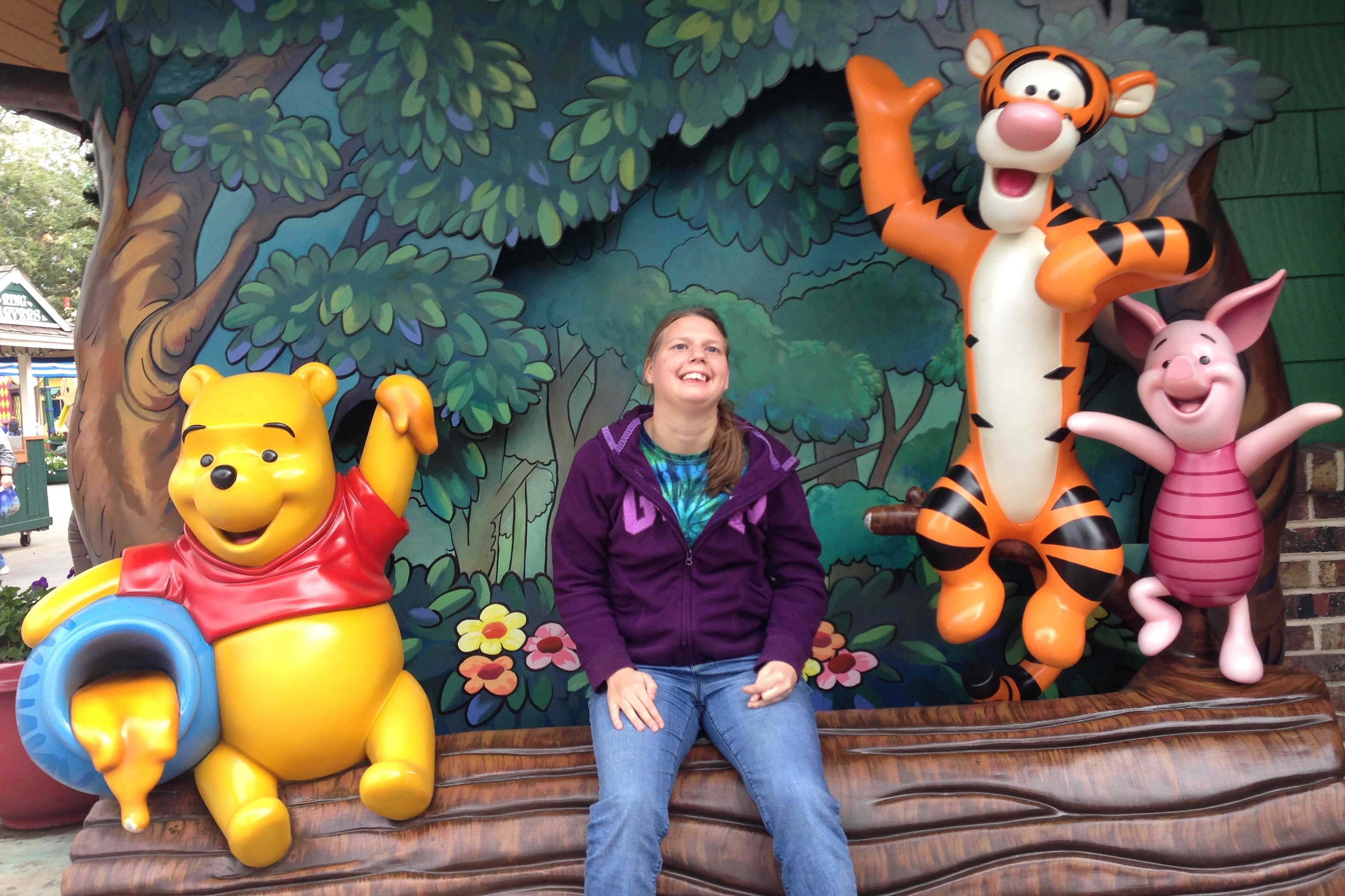 Smiling woman sitting on a bench with life-sized Winnie the Pooh, Tigger, and Piglet statues in front of a colorful forest backdrop at a theme park.