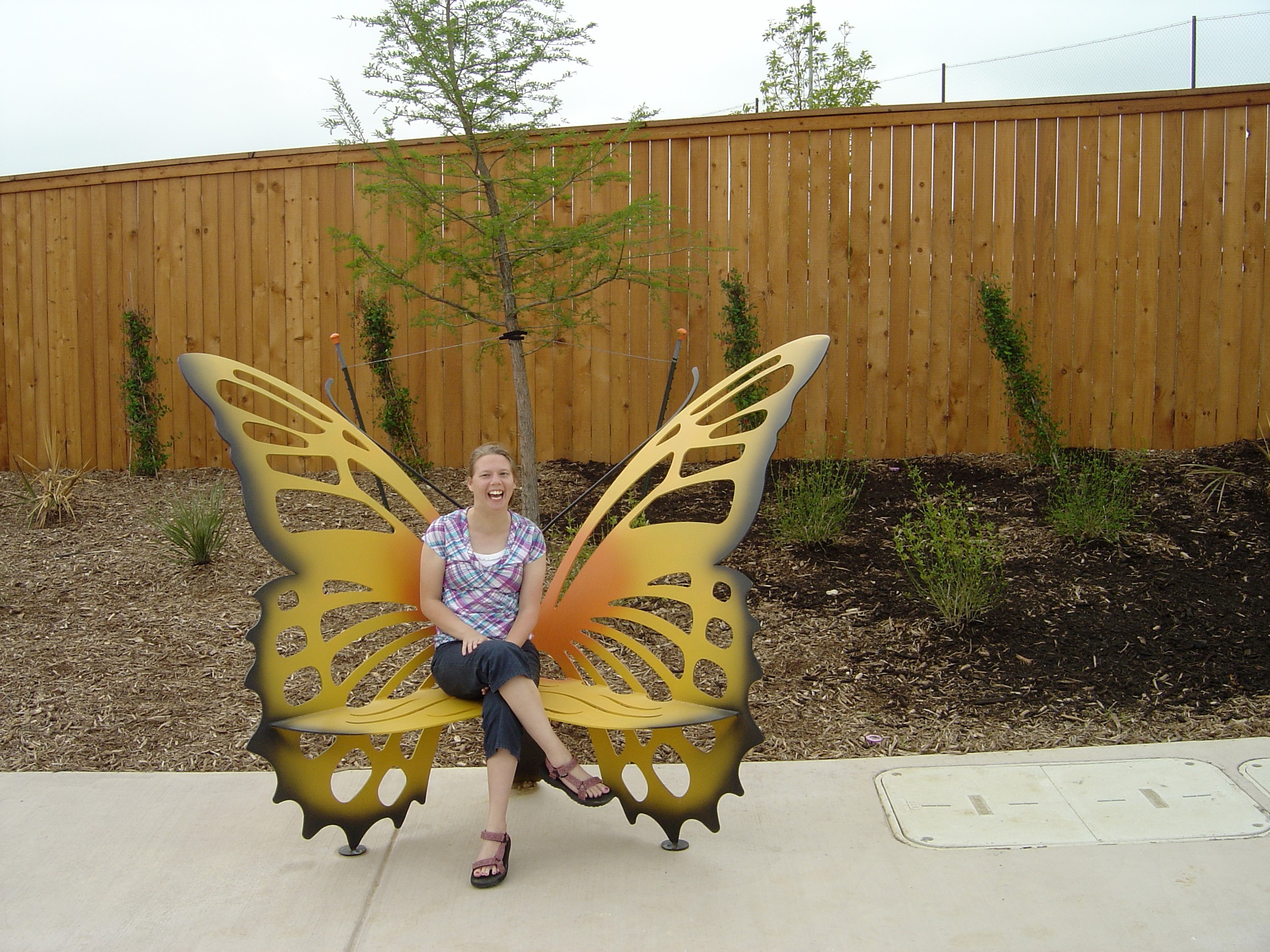 Smiling woman sitting on a decorative butterfly-shaped bench in an outdoor garden area with a wooden fence and young trees in the background.