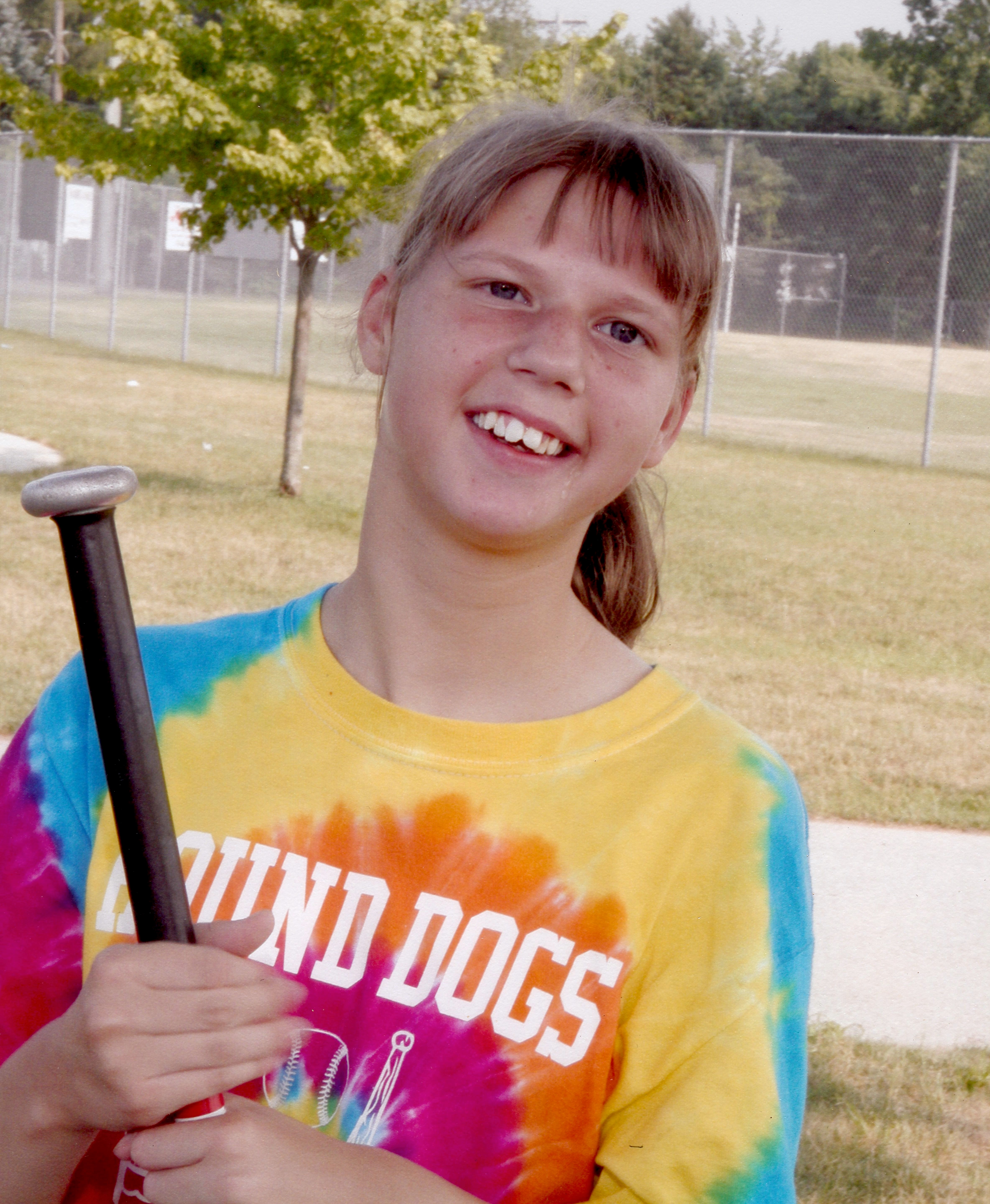 Smiling girl in a colorful tie-dye shirt holding a baseball bat at a park with trees and a chain-link fence in the background.
