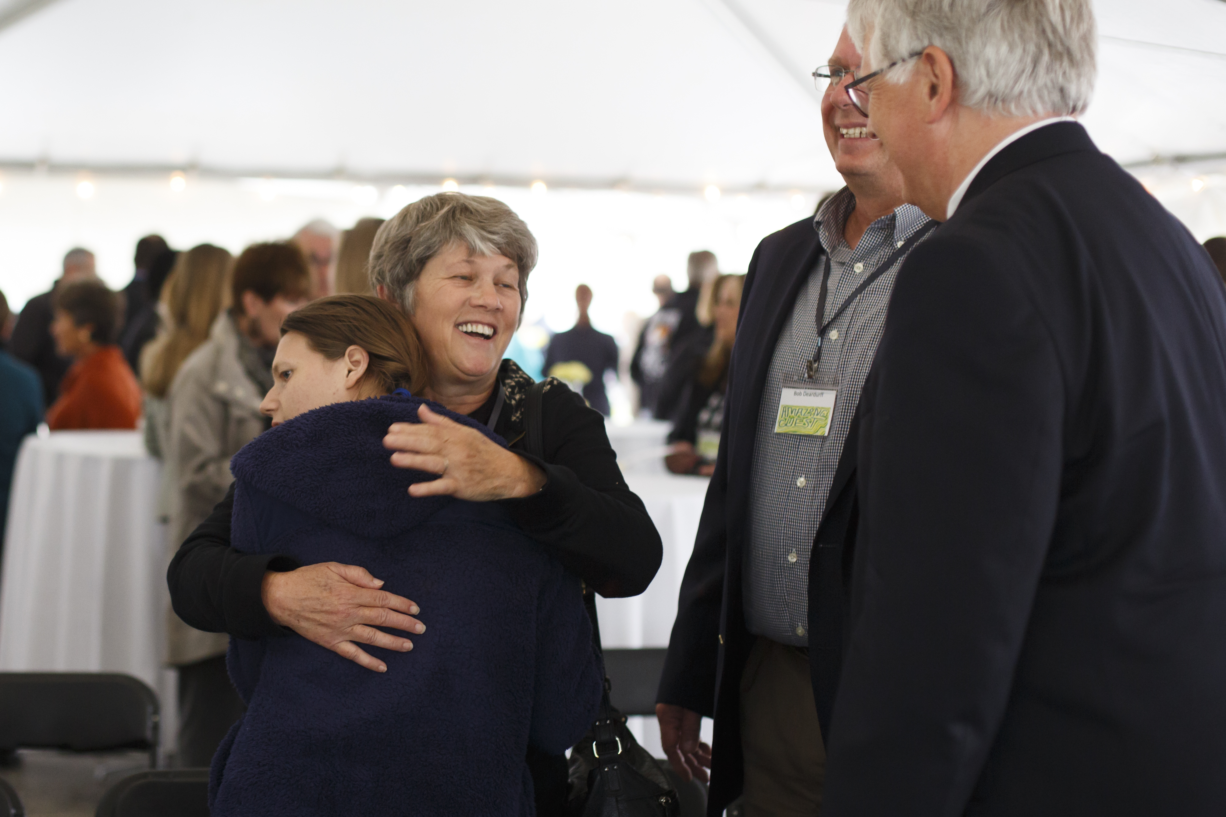 Smiling woman hugging a young woman in a blue jacket at an indoor event, surrounded by people socializing in the background.