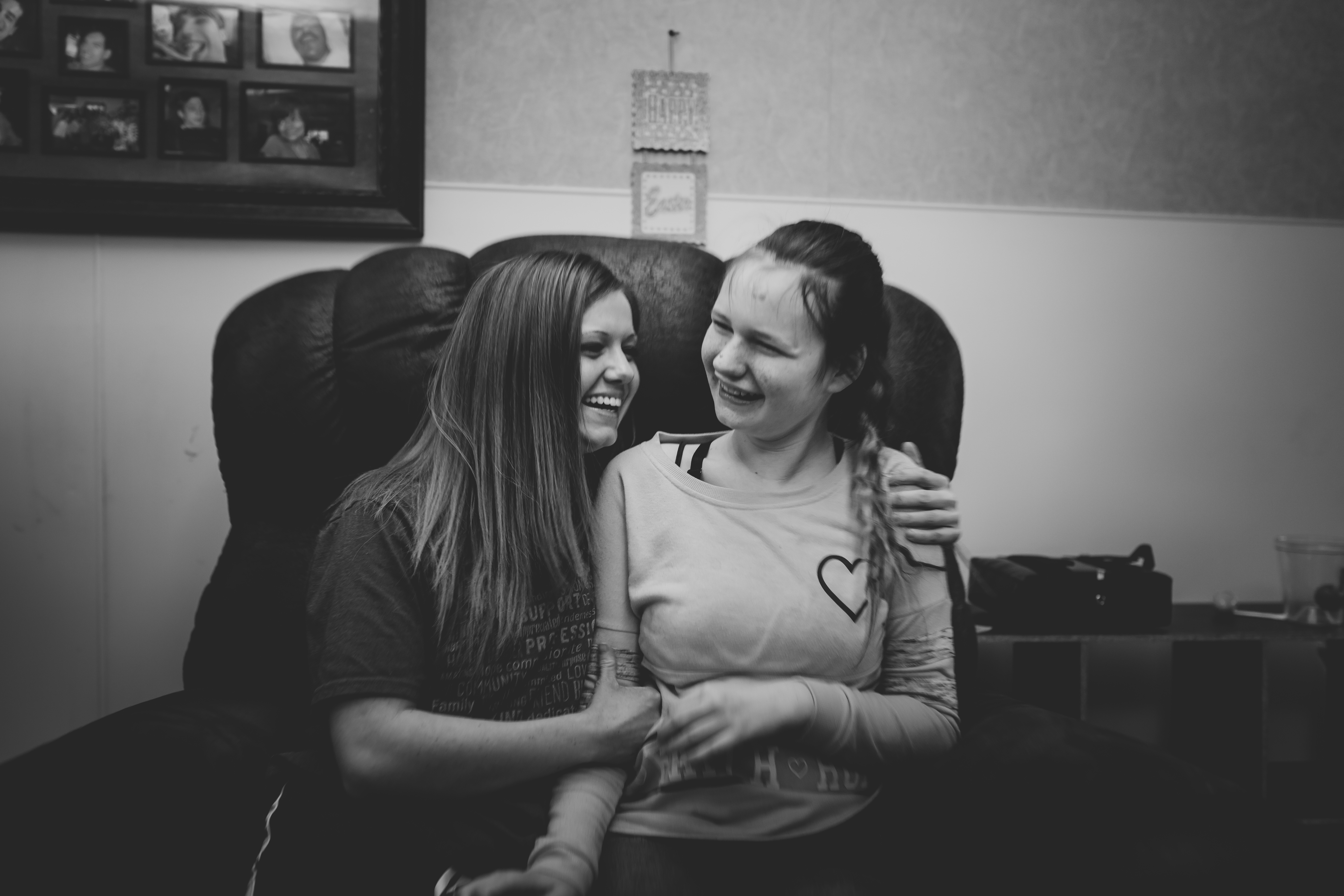 Black and white photo of two young women sitting together, laughing and sharing a happy moment on a chair indoors.
