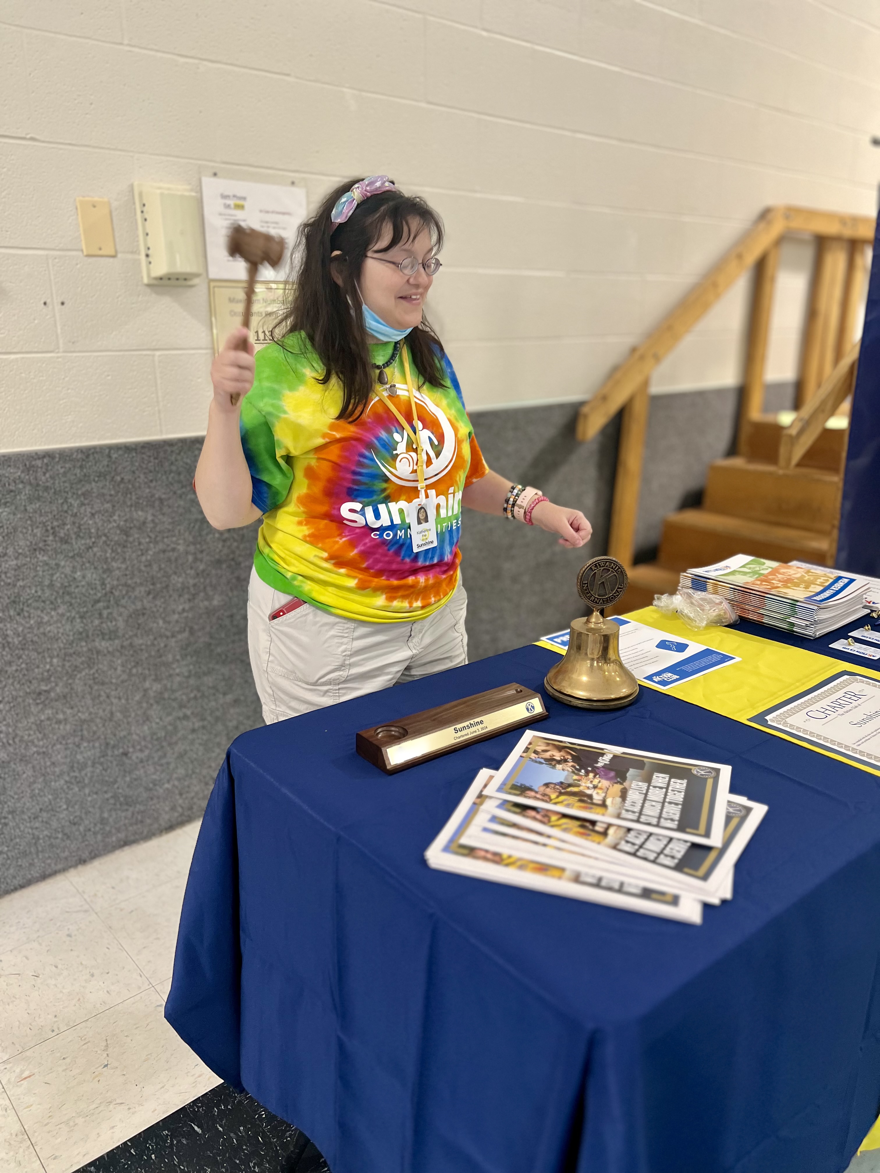 A Sunshine Aktion Club member in a colorful tie-dye Sunshine Communities shirt stands at a table holding a gavel, preparing to lead a meeting during the official charter ceremony in Maumee, Ohio. The table is set with Kiwanis materials, a bell, and a nameplate celebrating the new club.