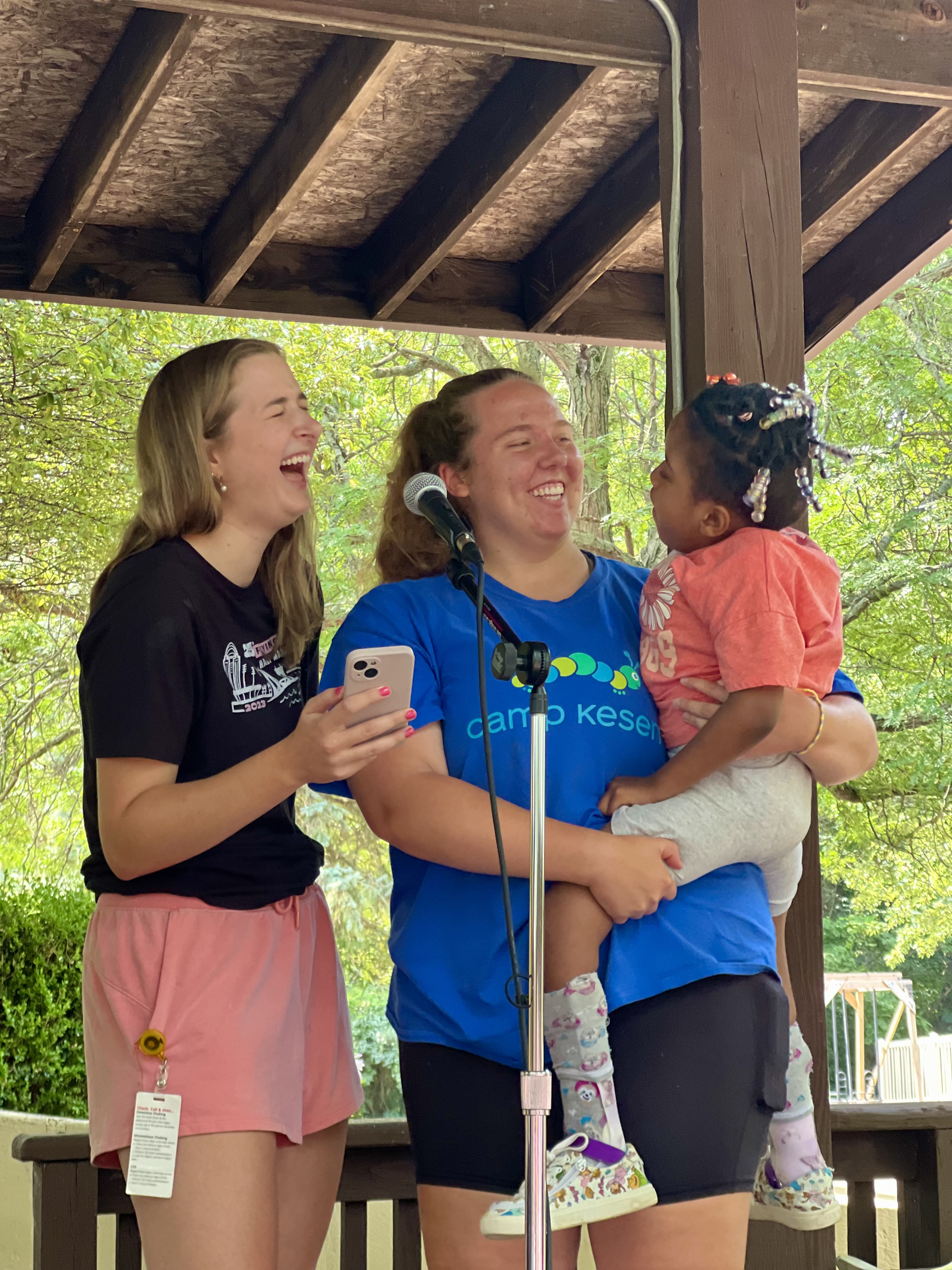 Two young women laughing together on stage at an outdoor camp event, one holding a toddler with beaded braids, near a microphone under a wooden pavilion.