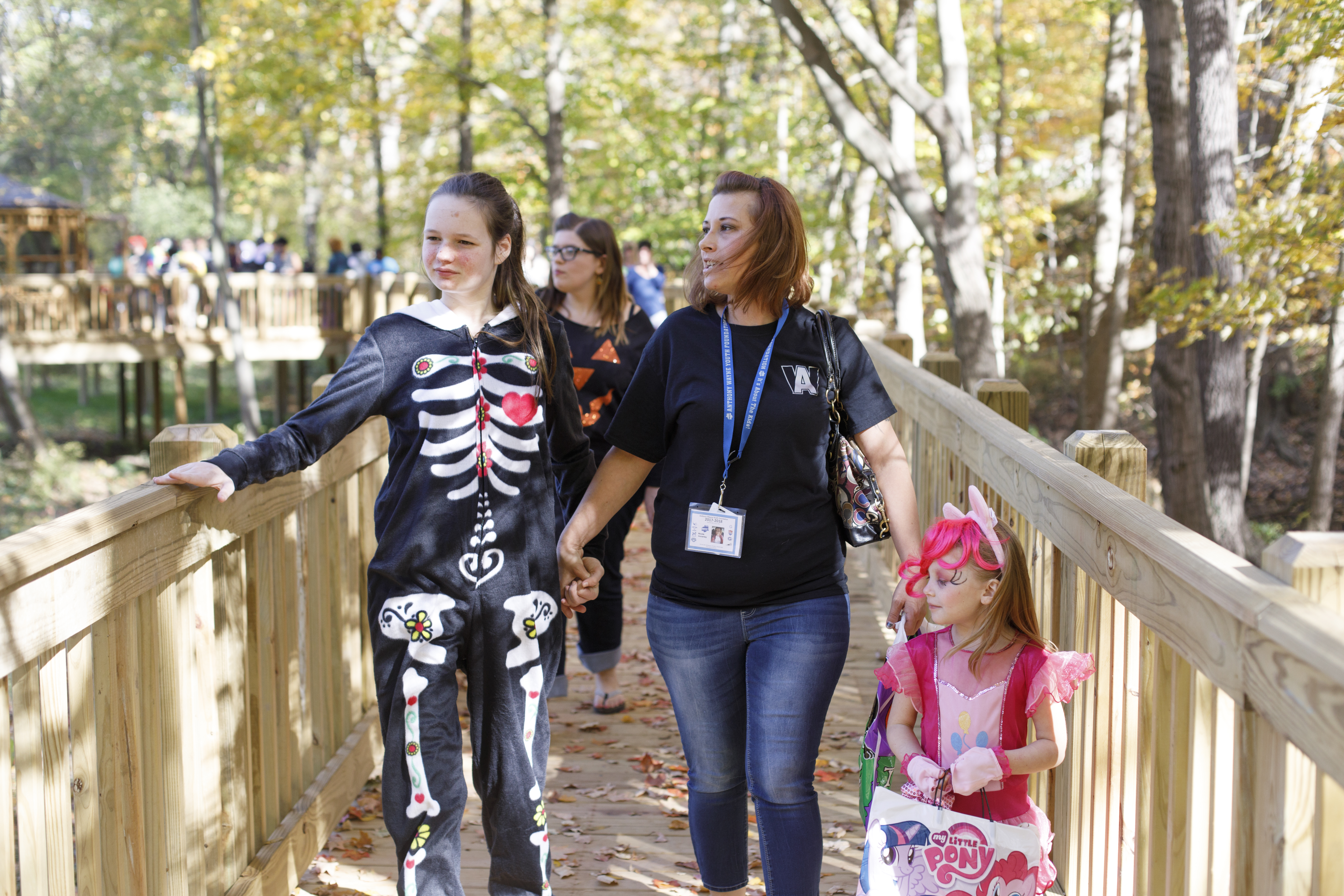 Woman walking with two girls in Halloween costumes on a wooden bridge in the forest during autumn.