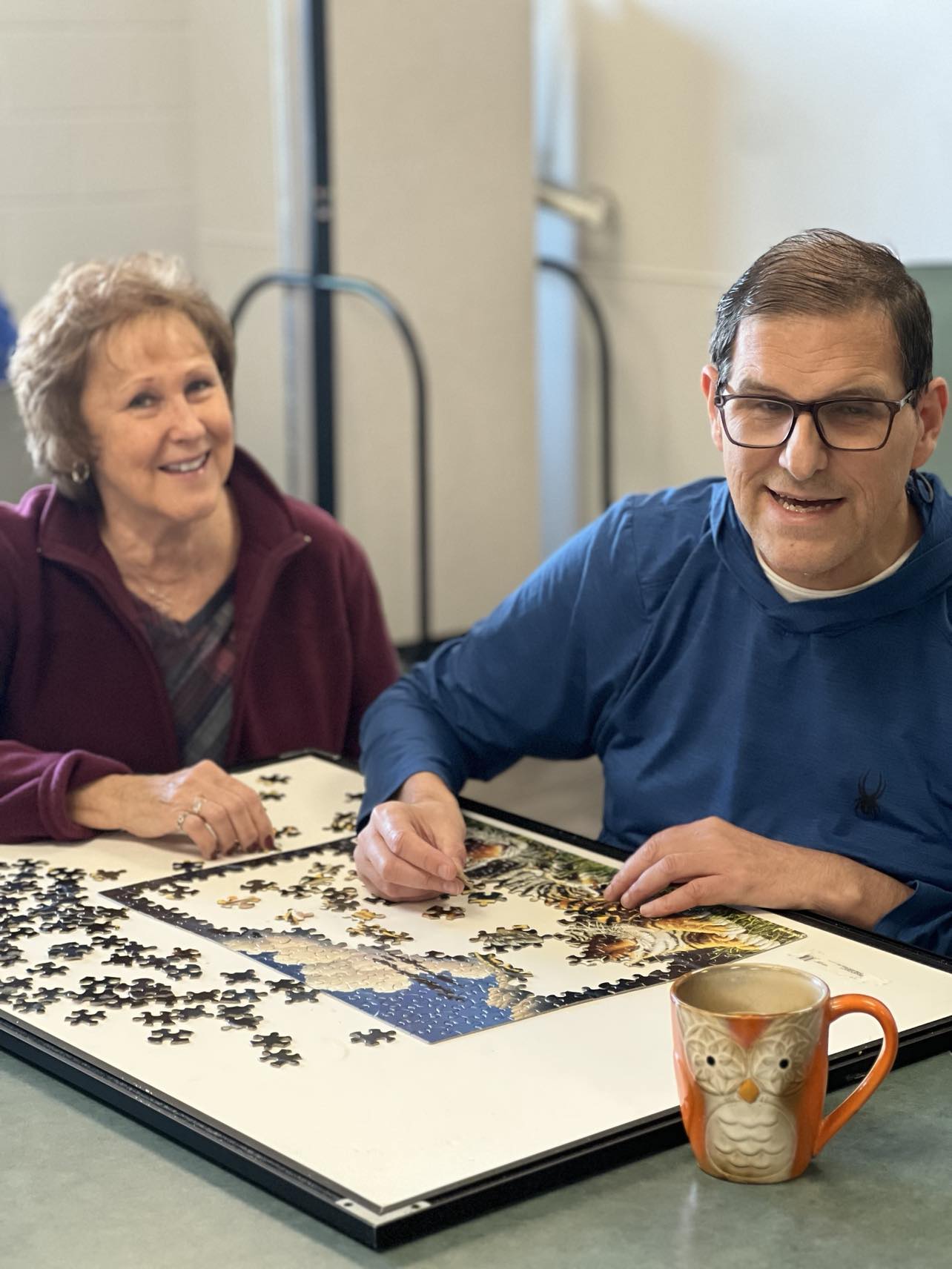"A smiling woman and man sit together working on a jigsaw puzzle. The woman, wearing a burgundy jacket, looks at the camera while the man, in glasses and a blue sweatshirt, holds a puzzle piece. A nearly completed puzzle with a tiger image is in front of them on the table, along with a ceramic owl mug."
