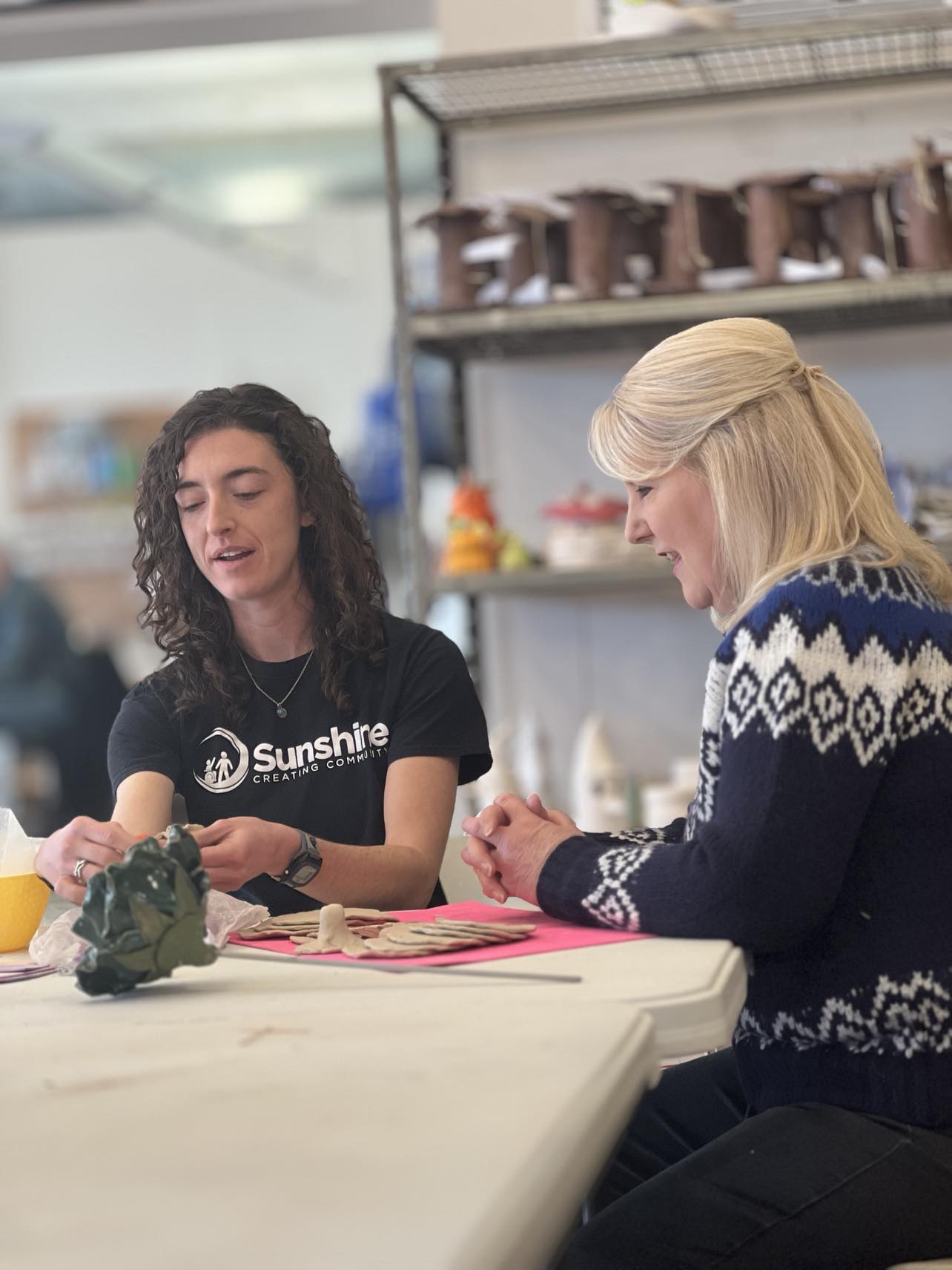 Marilyn Yakumithis, Studio Manager at Sunshine Studios, sits at a table with WTOL 11’s Lissa Guyton, guiding a hands-on clay art activity. The two women are engaged in conversation while working on ceramic pieces. Marilyn wears a black “Sunshine Creating Community” t-shirt, and Lissa is dressed in a navy blue and white sweater. Shelves filled with ceramic mushroom sculptures are visible in the background, highlighting the creative environment of the inclusive Sunshine Studios art program in Maumee, Ohio.