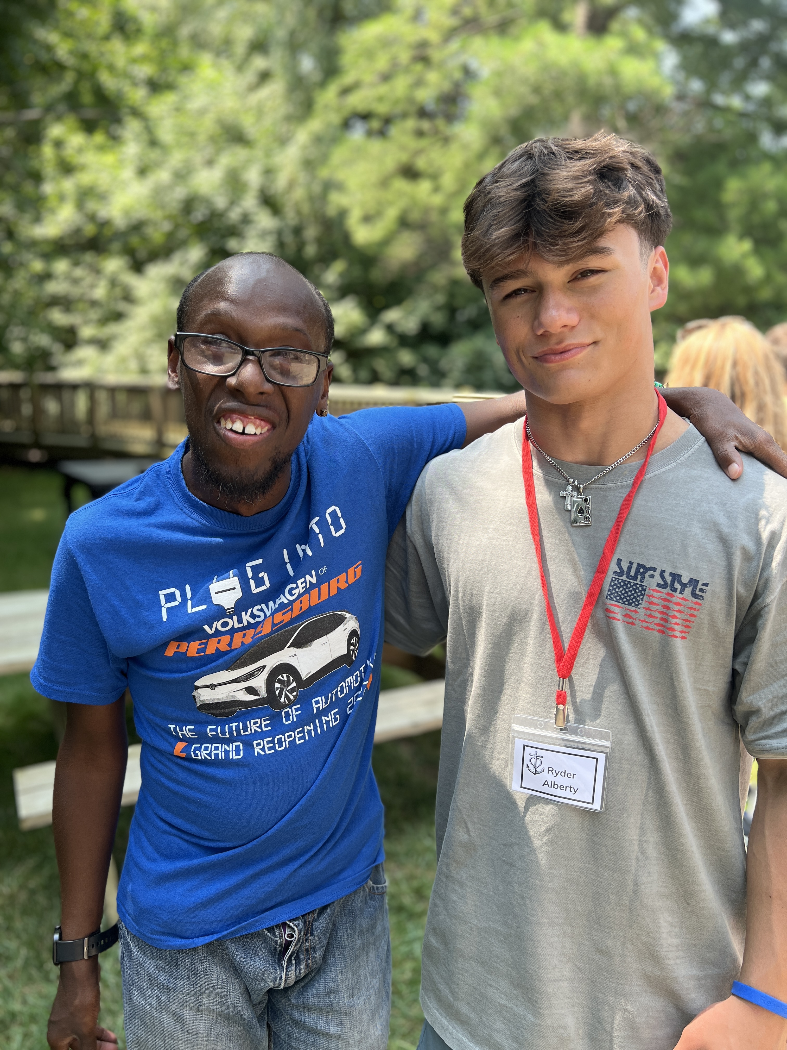 Two young men smiling side-by-side outside, one wearing a blue Volkswagen Perrysburg t-shirt and glasses, the other wearing a gray Sunshine shirt with a red lanyard and name tag reading "Ryder Alberty." Trees and a boardwalk are in the background.