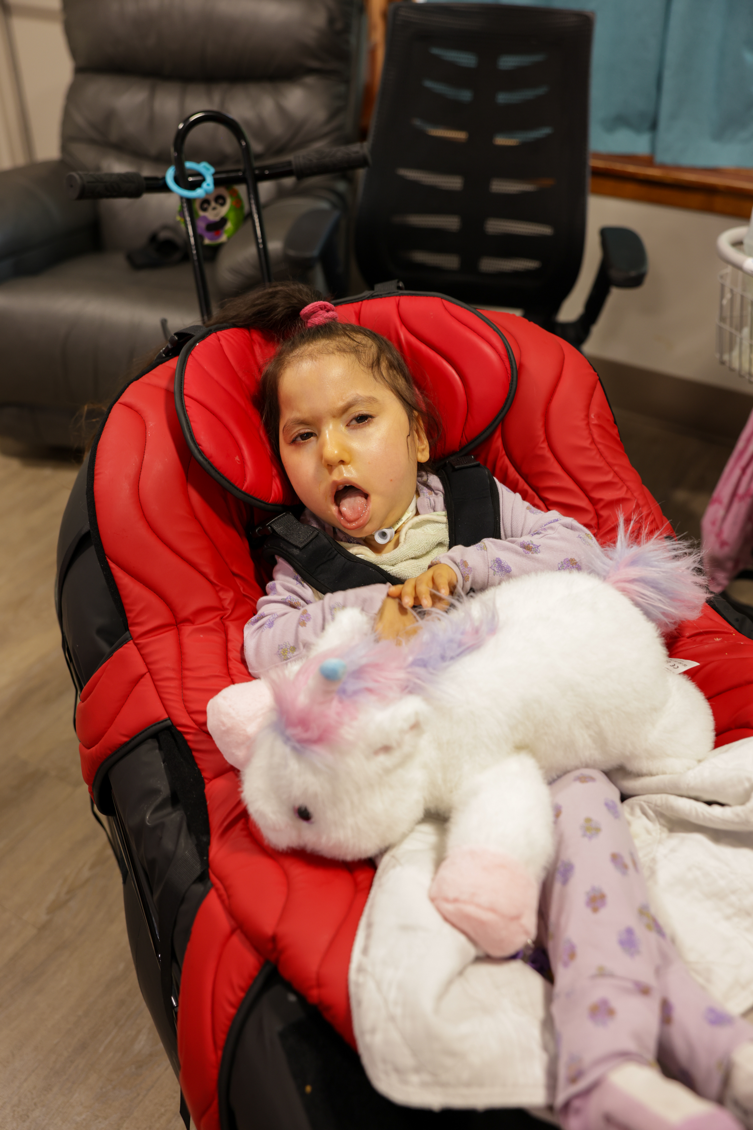 A young child lies comfortably in a padded red medical chair, supported with a harness. The child has a tracheostomy tube and rests with eyes partially open, tongue slightly out.