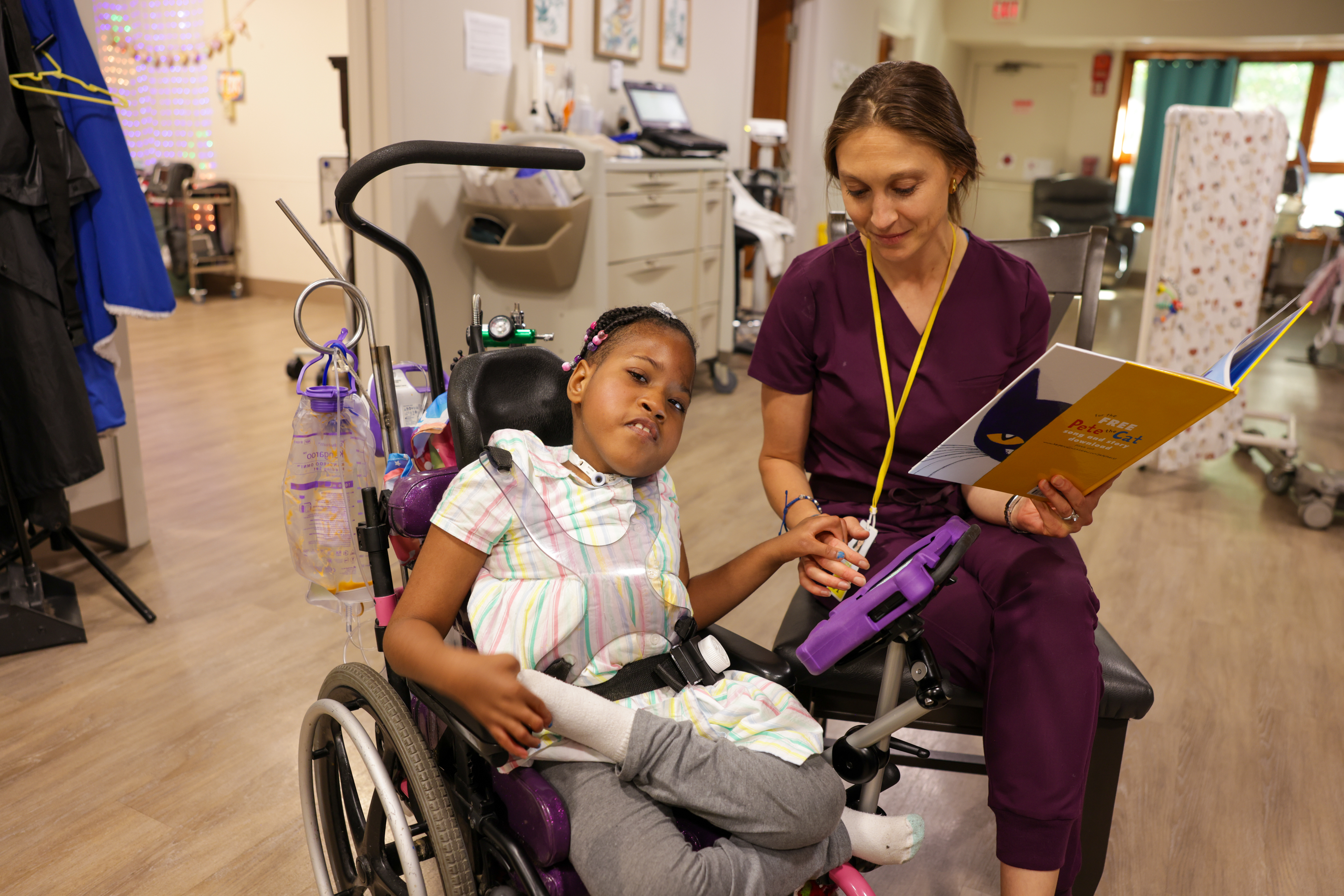A young girl sits in a wheelchair with medical equipment attached, wearing a colorful striped dress and gray pants. She holds hands with a woman in purple scrubs who is seated beside her, reading from a children’s book titled “Pete the Cat.”