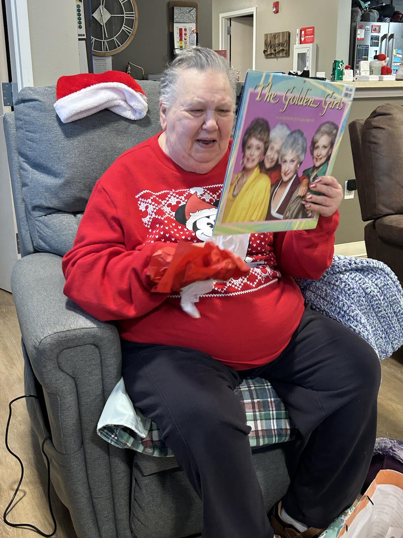 Elderly woman in a Mickey Mouse Christmas sweater joyfully opens a gift and holds a Golden Girls record, celebrating the holidays in a cozy group home setting.