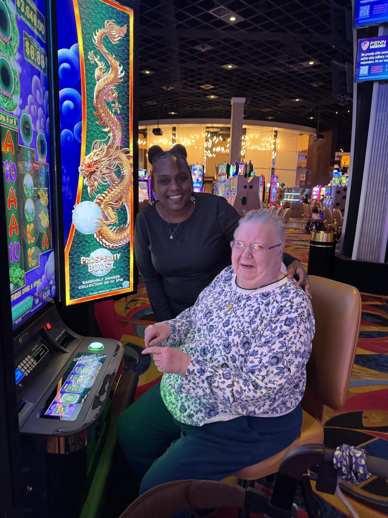 Two women enjoy time at a casino, one smiling while seated at a slot machine and the other standing beside her, showcasing inclusive community outings supported by Sunshine Communities.