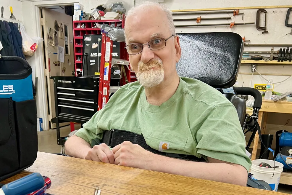 "A man with light skin, short white hair, and glasses sits in a wheelchair, wearing a light green shirt. He is in a workshop setting with various tools and hardware neatly organized in trays on the table in front of him. The background shows shelves with more tools and workshop equipment. The man is smiling slightly, and the environment suggests he is engaged in sorting or working with tools."