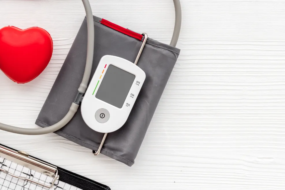 The image shows a blood pressure monitor with a cuff, a red heart-shaped object, and a clipboard with an electrocardiogram (ECG) graph, all placed on a white wooden surface. Let me know if you need any modifications or descriptions for specific use!