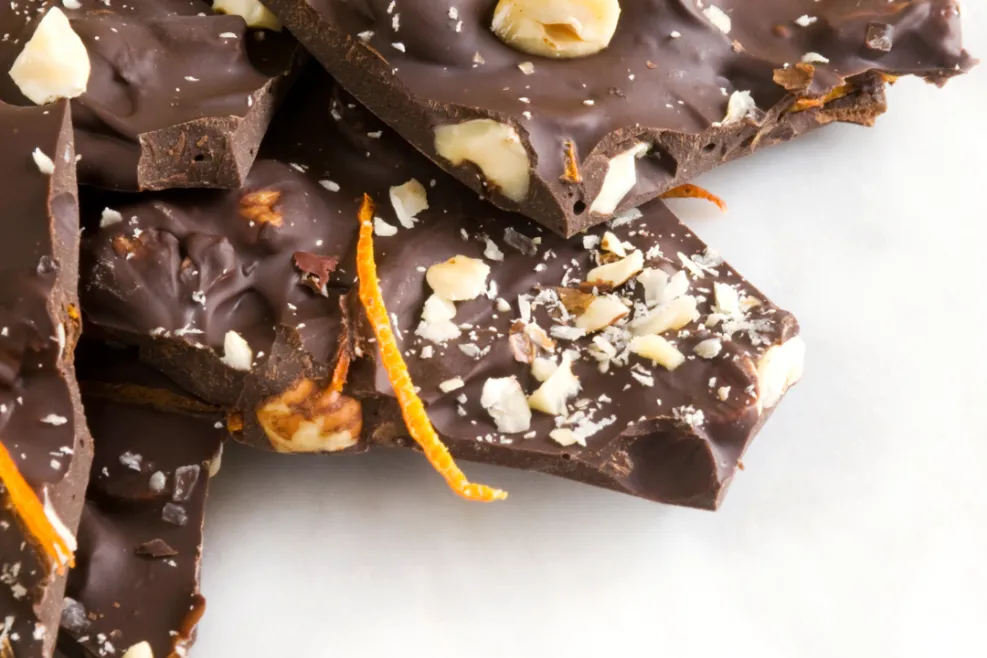 "Close-up of dark chocolate bark with hazelnuts, orange zest, and white chocolate shavings on a marble surface. The bark is broken into irregular pieces, showcasing a rich, glossy texture with crunchy nut and citrus accents."