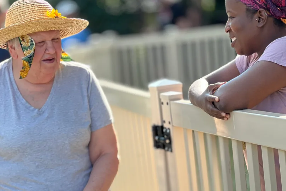 "An elderly woman with light skin, wearing a straw hat adorned with a yellow flower and patterned scarf, walks with a cane while engaging in conversation with a younger woman with dark skin, who is leaning on a beige fence. The younger woman wears a colorful headwrap and a pink shirt, smiling warmly as they talk. The background is blurred with trees and other people visible in the distance, suggesting an outdoor setting."