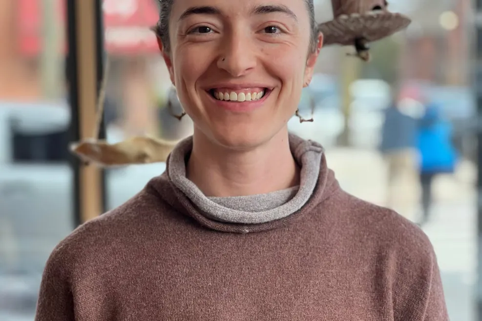  A woman with dark hair pulled back smiles warmly at the camera. She is wearing a brown sweater layered over a gray cowl-neck shirt. Behind her, there is a large window with a view of a street and some hanging natural sculptures or art pieces, including a branch with a carved wooden object. The setting appears to be an art studio or café with a cozy, creative vibe.