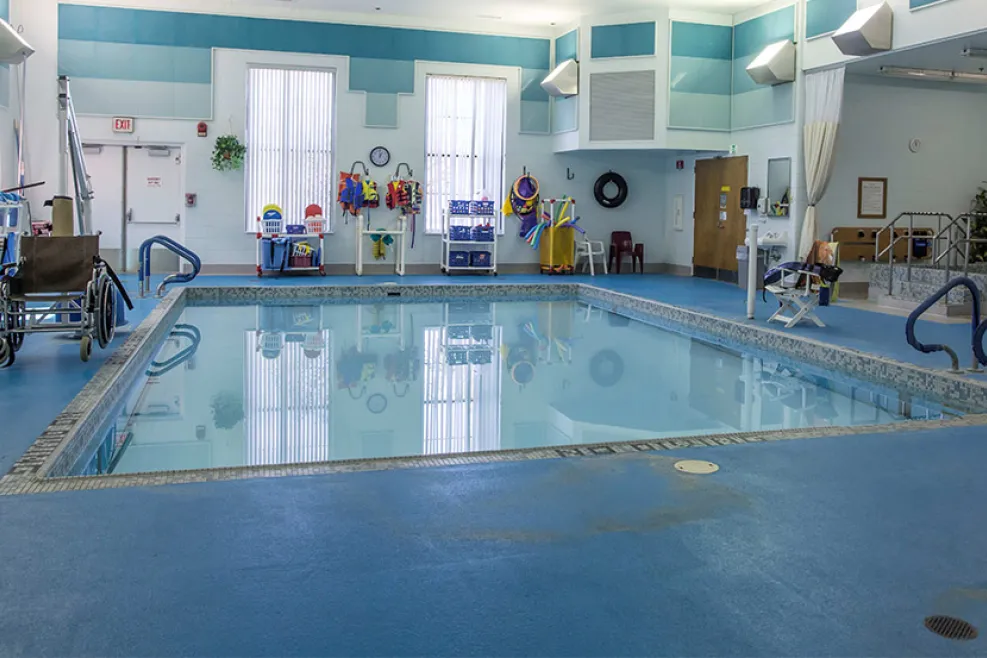 "Indoor therapy pool surrounded by blue flooring and equipped with accessibility features, including a wheelchair lift and pool chairs. Colorful life jackets, pool noodles, and safety equipment are organized along the far wall."