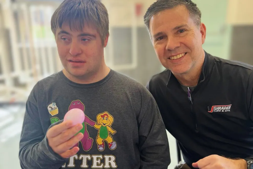 A man from Shrader Tire & Oil smiles beside a young adult with Down syndrome as they participate in an Easter-themed volunteer activity at Sunshine Communities in Maumee, Ohio. The young adult holds a dyed egg, wearing a “Better Together” shirt, with colorful cups of dye in the foreground.