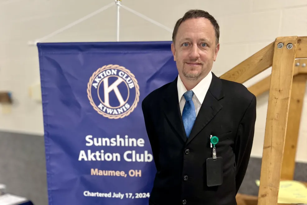 A Sunshine Aktion Club member in a black suit and blue tie stands proudly next to a Kiwanis banner at the Sunshine Communities charter event in Maumee, Ohio, July 17, 2024.