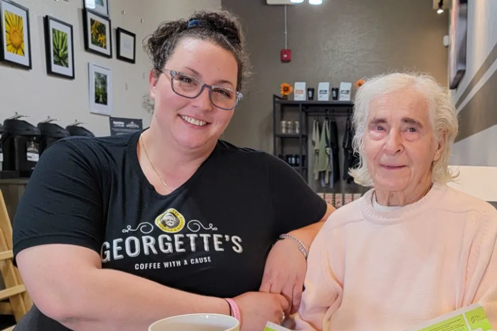 Smiling woman in a Georgette’s Coffee with a Cause t-shirt sitting beside an elderly woman at Georgette’s café in Maumee, Ohio, enjoying coffee and community.