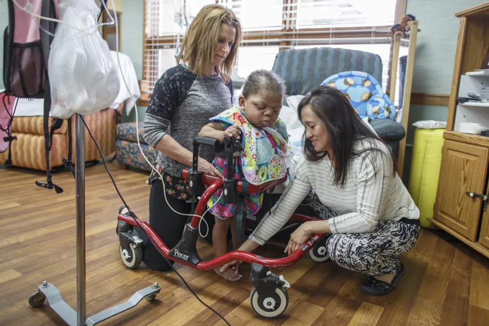 Physical therapists assist a young child using a red walker during a therapy session in a rehabilitation center with supportive equipment and caring staff.