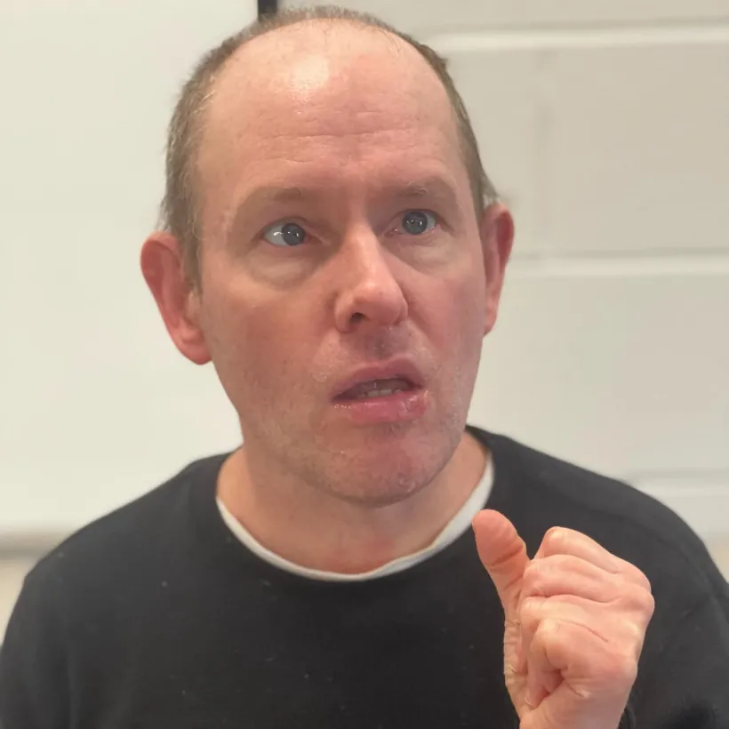 A man with short, light brown hair and blue eyes is wearing a black sweater. He has an expressive facial expression and is gesturing with his right hand, appearing engaged in conversation. The background features a white brick wall.