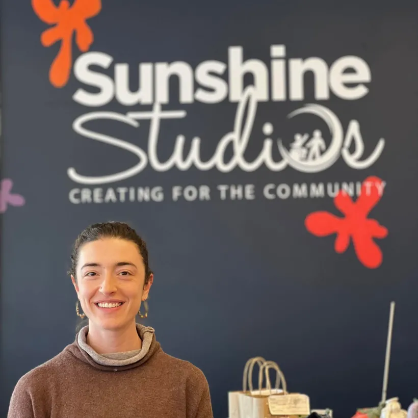 Marilyn Yakumithis, new manager of Sunshine Studios in Maumee, Ohio, stands smiling in front of the studio’s branded wall featuring the phrase “Creating for the Community.” A professional artist with international experience, Marilyn leads the inclusive art studio that supports individuals with developmental disabilities and showcases locally crafted ceramic pieces and handmade goods.
