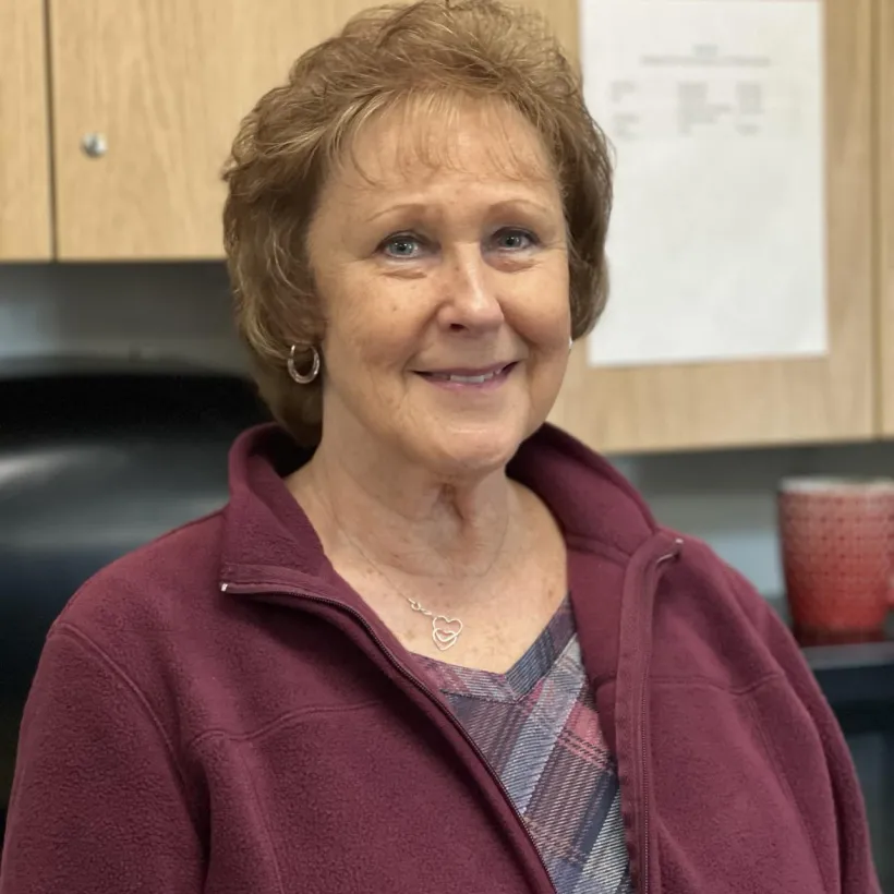 "Smiling woman with short, curly light brown hair wearing a maroon fleece jacket and plaid top, standing in front of wooden cabinets in an indoor setting. A red cup and paper on the cabinet are visible in the background."