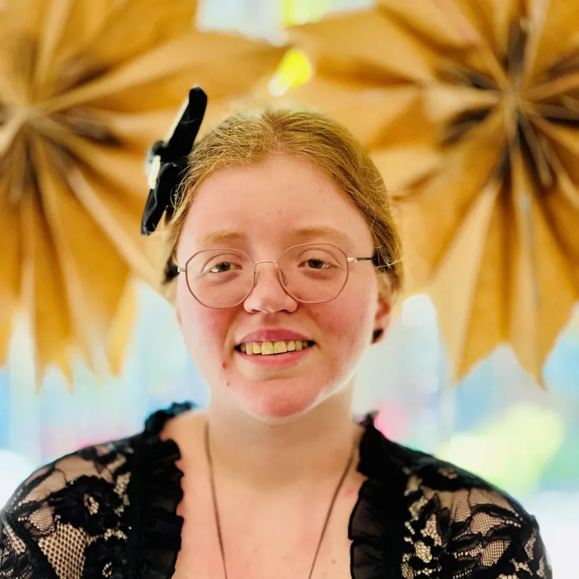 Portrait of Brooke, a Sunshine Studios artist, wearing glasses and a black lace top with a bow in her hair. She stands smiling in front of a vibrant background decorated with large paper starbursts, representing her creative spirit and quiet confidence.