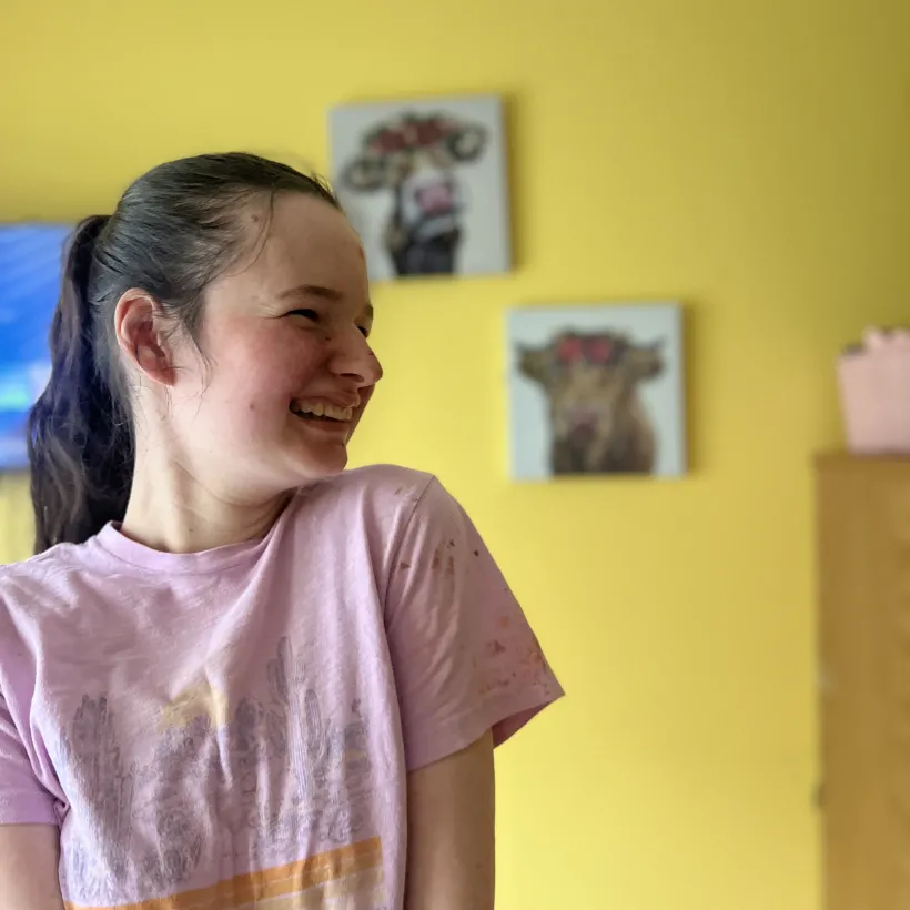 Smiling young girl in a pink t-shirt standing against a bright yellow wall with playful animal artwork in the background.