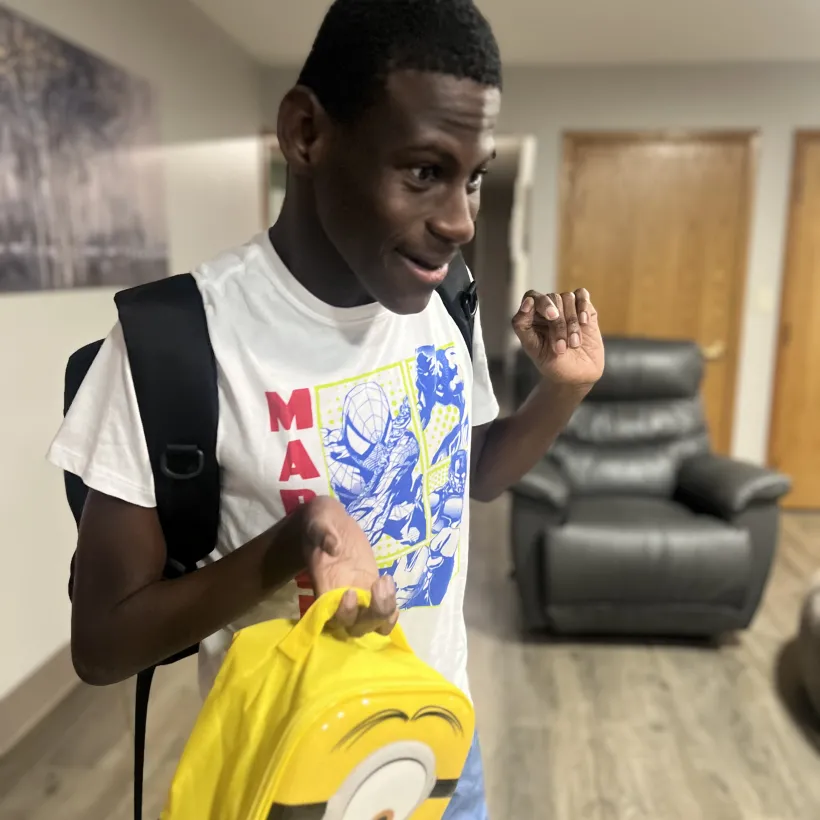 Young man smiling indoors, wearing a Marvel T-shirt and backpack, holding a bright yellow Minions-themed bag.