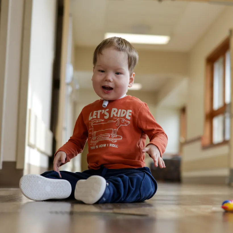 A young child sits on the floor of a bright hallway with large windows