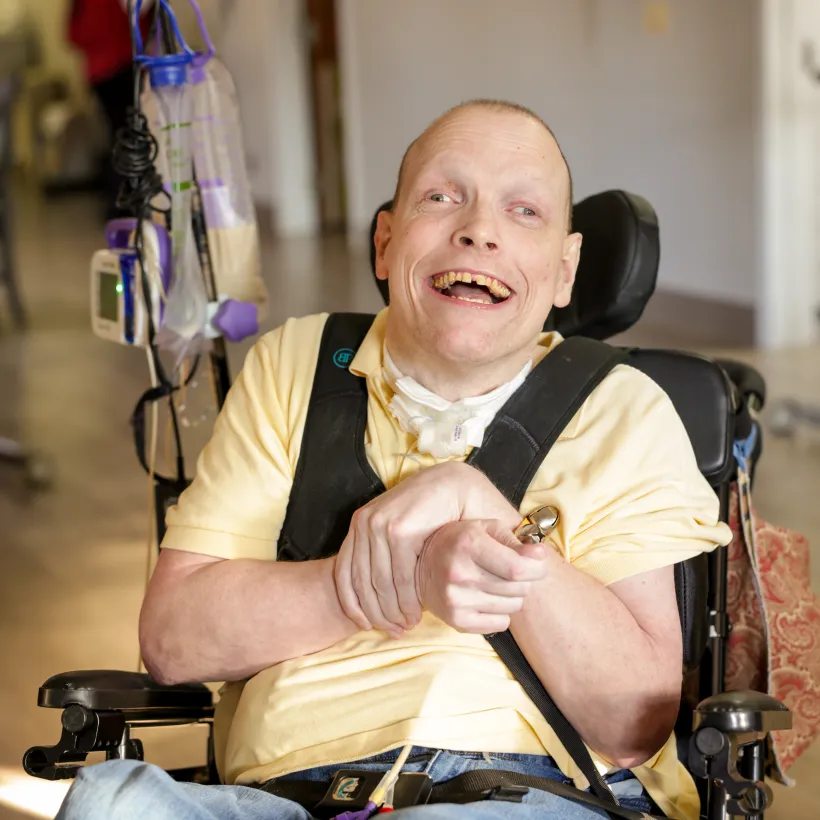 A smiling adult man sits in a motorized wheelchair in a bright indoor hallway.