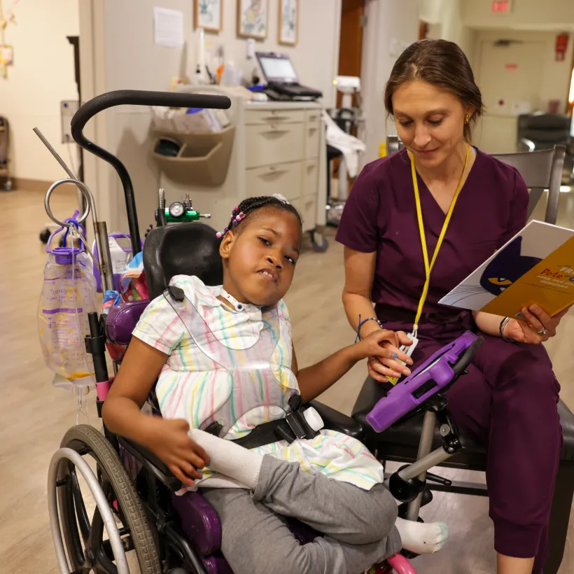 A young girl sits in a wheelchair with medical equipment attached, wearing a colorful striped dress and gray pants. She holds hands with a woman in purple scrubs who is seated beside her, reading from a children’s book titled “Pete the Cat.”